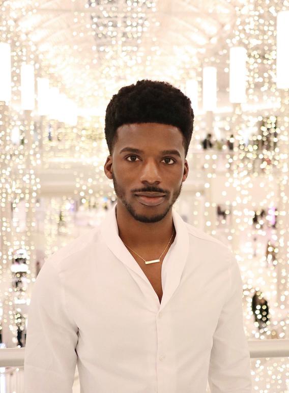 A Black man looks at the camera while wearing white and standing in front of string lights in a large venue.