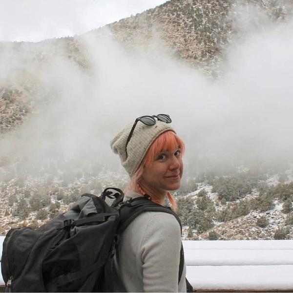 Image of a young woman with pink hair in a light grey stocking hat (with sunglasses on the hat) wearing a grey long sleeve shirt and a big, full black backpack. She is looking at the camera, but behind her we can see a mountain valley filled with scrub brush and a lot of mist/fog.