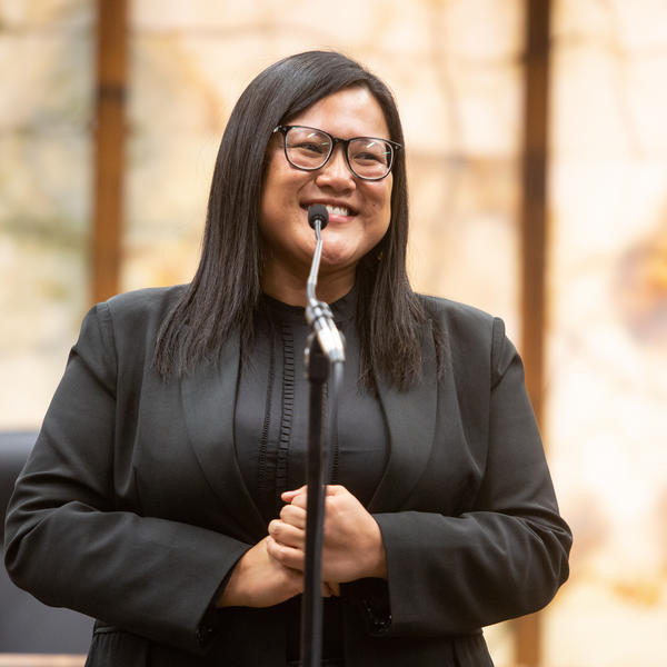 Woman with dark brown hair wearing glasses and a black blazer standing in front of a microphone