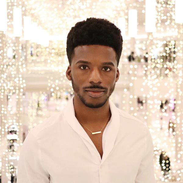 A Black man looks at the camera while wearing white and standing in front of string lights in a large venue.