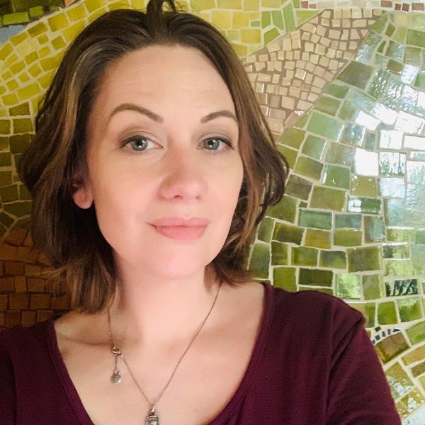 A white woman with brown hair wearing a burgundy shirt stands in front of a green and yellow mosaic tile wall
