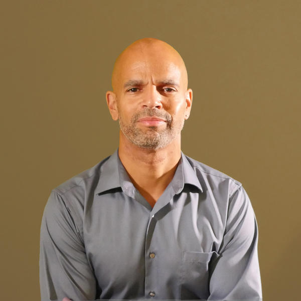 A light skin Black man wearing a gray button down standing in front of a tan background