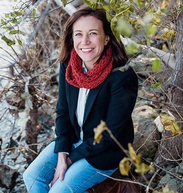 White woman with brown hair smiling wearing a red scarf, a white button up and a dark blue blazer and jeans sitting in a forest.