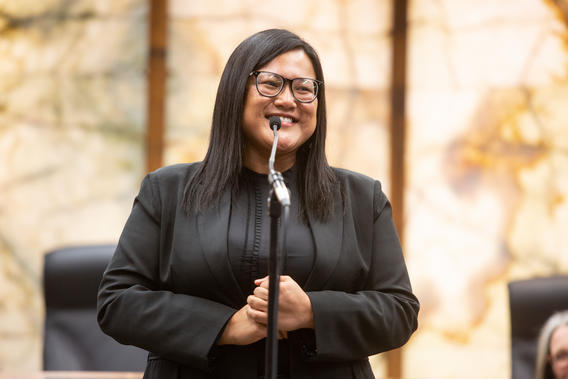 Woman with dark brown hair wearing glasses and a black blazer standing in front of a microphone