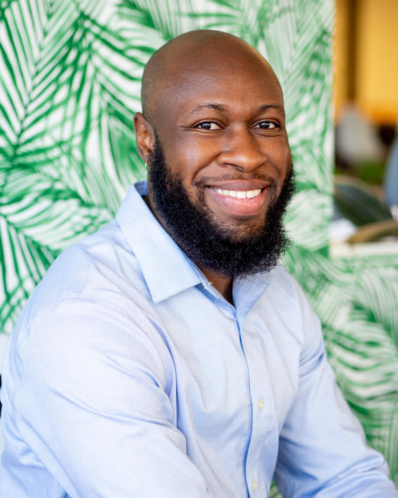 Black man with a beard smiles while wearing a blue long sleeved shirt and standing in front of a green palm leaf patterned wall.