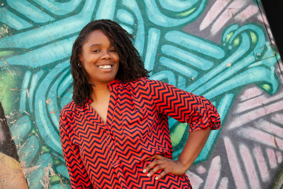 A Black woman wearing a red and black zig-zag patterned top smiles with her hand on her hip while standing in front of a turquoise and white mural.