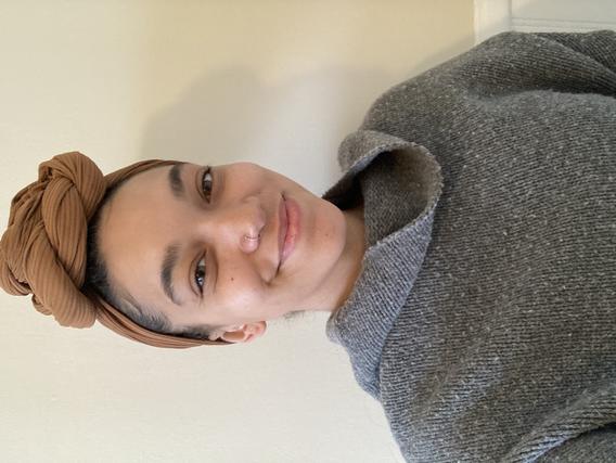 A Black woman smiling wearing a mustard colored head wrap and a gray sweater.