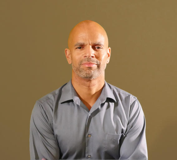 A light skin Black man wearing a gray button down standing in front of a tan background