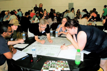 A group of 5 people huddled around a table with a map or a chart of some sort. You can see other similar groups in the background.