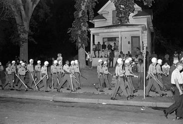 black and white photograph of large group of uniformed police marching passed a house during the nighttime