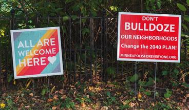two lawn signs side-by-side. Sign on left reads "All Are Welcome Here". Sign on the right reads "Don't Bulldoze Our Neighborhoods, Change the 2040 Plan"